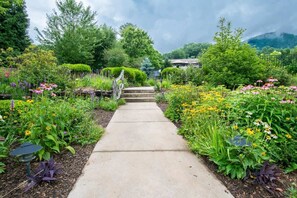 A garden pathway leading to a house with a thatched roof, surrounded by lush greenery and various flowering plants.