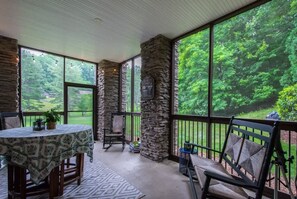 A covered patio with stone walls, a table with a patterned tablecloth, and chairs. - Scenic 2BR Condo w/ Porch + Pool Near Slope/Trails | Free Tickets (Maggie Valley)