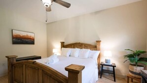 A bedroom with a wooden bed, two bedside tables with lamps, a ceiling fan, a painting on the wall, and a potted plant.