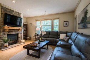 A living room with a stone fireplace, a leather sofa, a flat-screen TV, and a large window with blinds. - Scenic 2BR Condo w/ Porch + Pool Near Slope/Trails | Free Tickets (Maggie Valley)