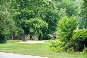 A lush green golf course with a stone bridge, surrounded by dense trees and well-maintained grass.