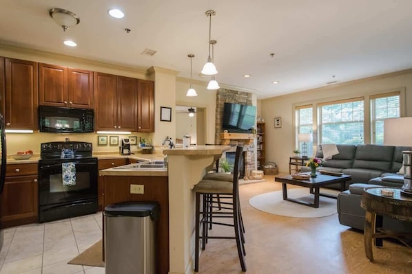 A modern kitchen with wooden cabinets, a microwave, and a stove. There is a dining area with a bar stool and a sofa.