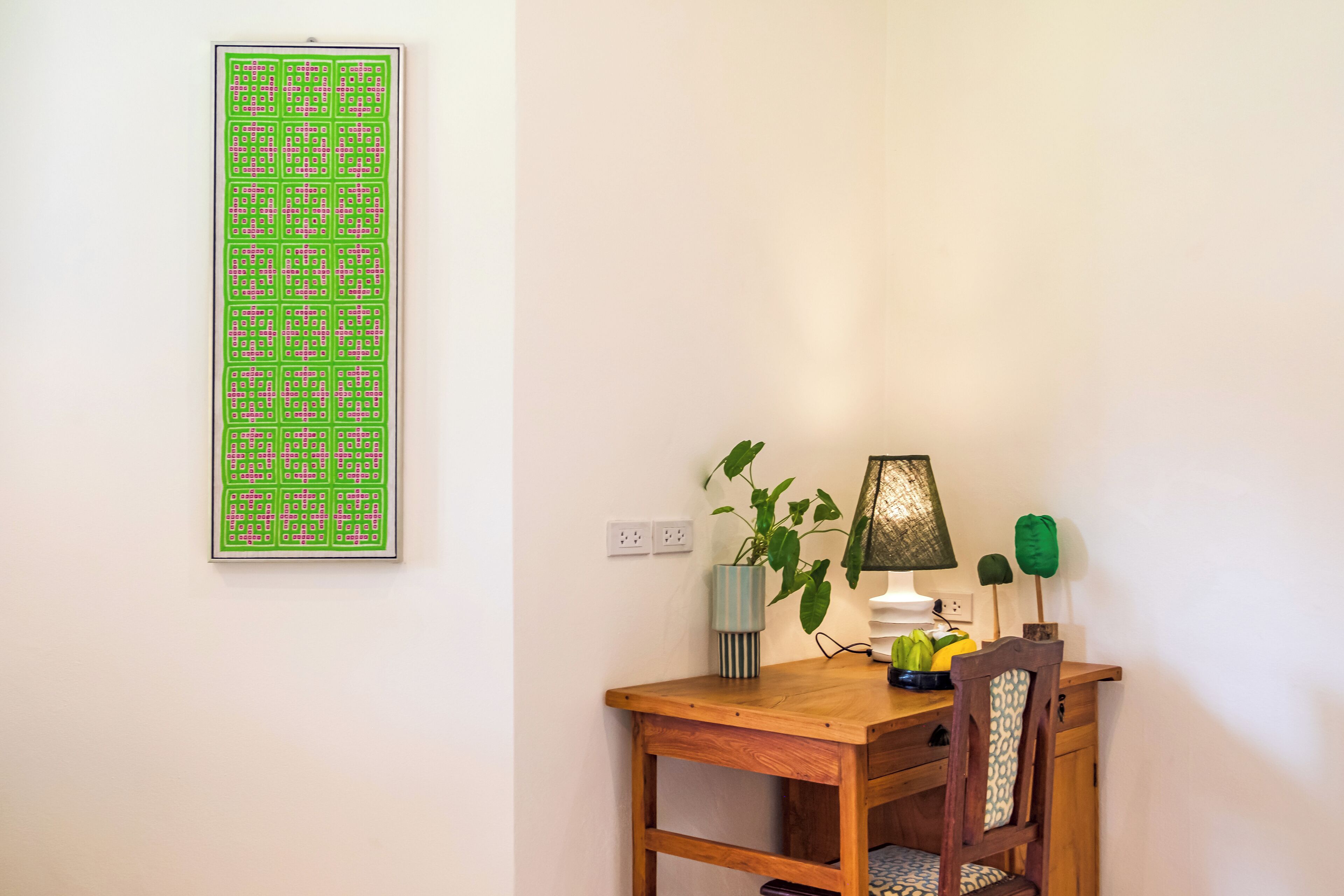 A wooden desk with a lamp, a plant, and a framed artwork on a white wall.