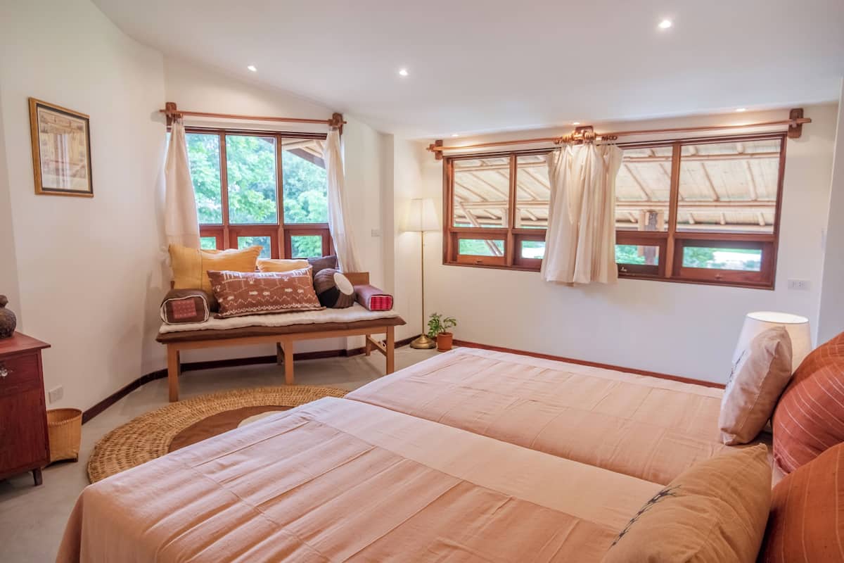 A bedroom with a large bed, a bench with cushions, a bedside table, a lamp, and windows with curtains.