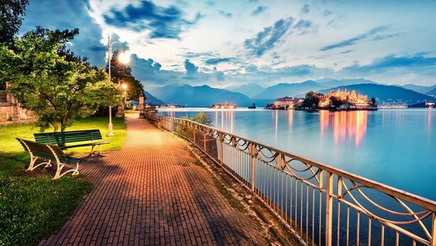 A waterfront promenade with a bench, a paved walkway, and a railing overlooking a calm lake with mountains in the background. - The Umbrella Cat - Lake Maggiore (Carpugnino)
