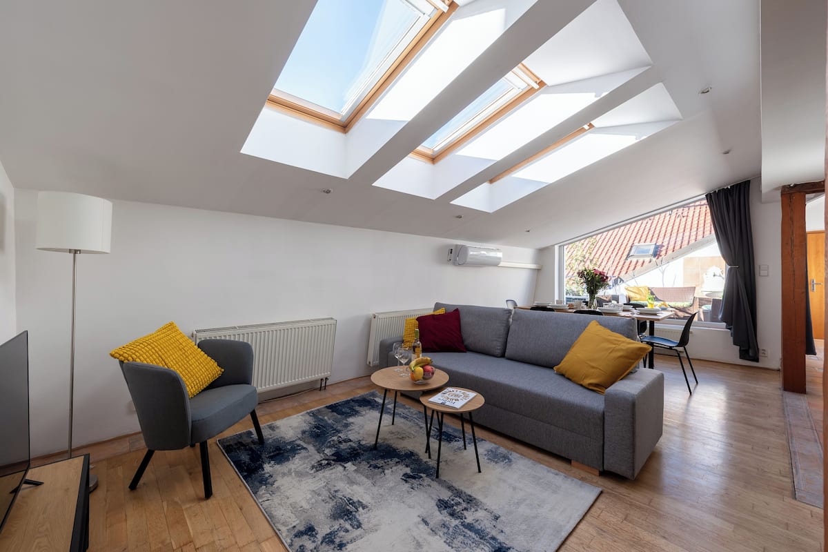 A modern living room with a grey sofa, a small round table, and a blue and white rug. There are skylights above the sofa, and a window with a view of the outside.