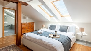 Une chambre avec une lucarne, un lit sur lequel se trouve un plateau avec des verres à vin et une bouteille, une commode et une table de chevet avec une lampe.