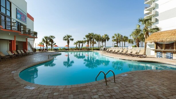 A pool area with lounge chairs, palm trees, and a building with a sign. - Resort View Two Room Condo Family-Friendly (Myrtle Beach)