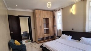 A bedroom with a large bed, a chair, a wardrobe, and a small table with an iron. - Starlight Homes (Kampala)