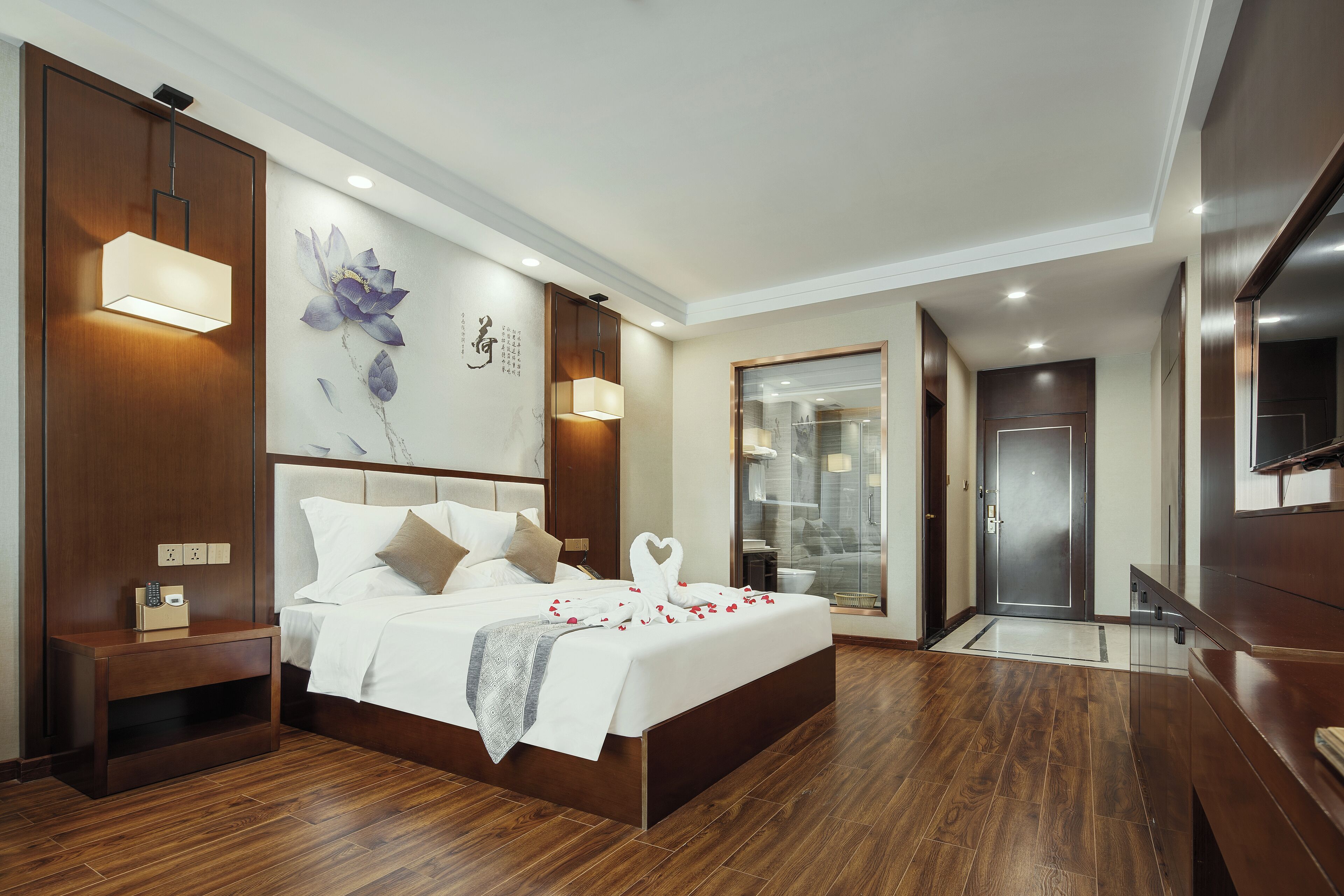 Une chambre d’hôtel moderne dotée d’un grand lit, d’un plancher en bois, d’une table de chevet, d’un téléviseur fixé au mur et d’une salle de bain visible à travers une porte en verre.