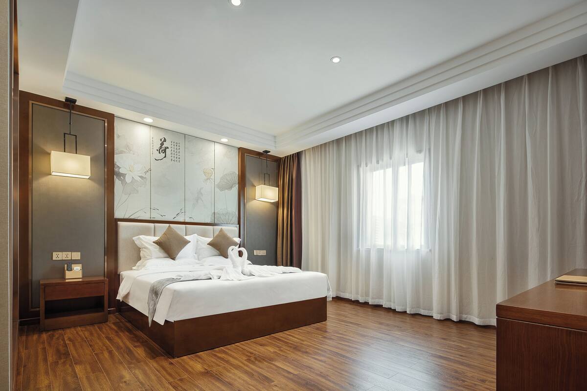 Une chambre d’hôtel avec un grand lit, une table de chevet avec une lampe, un plancher en bois et une grande fenêtre avec des rideaux.
