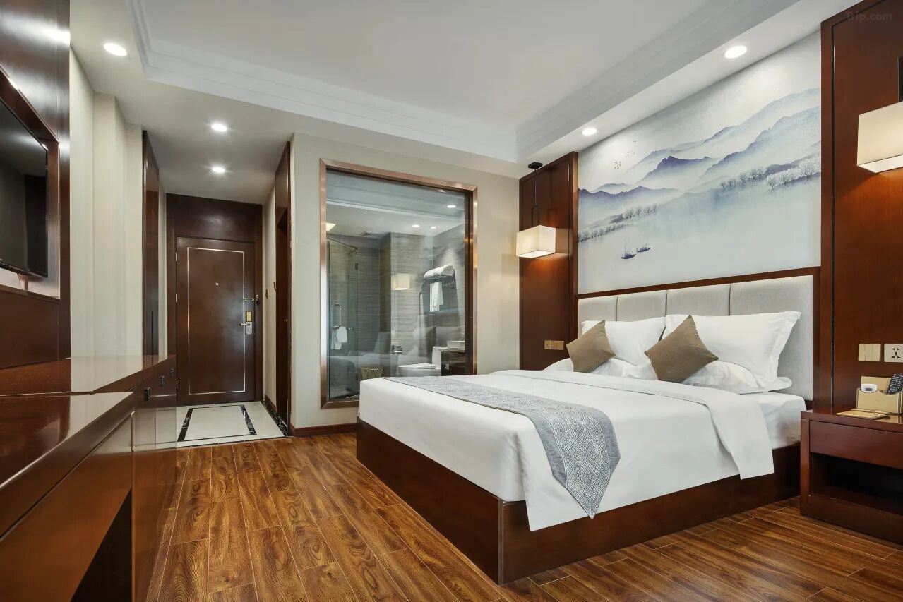 A modern hotel room with a wooden floor, a large bed with white bedding, a glass-enclosed shower, and a painting of mountains on the wall.