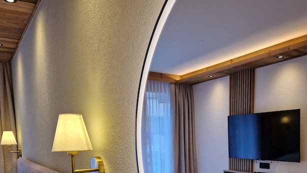 A modern hotel room with a large mirror, a bed with beige bedding, a wooden nightstand, a TV on the wall, and a window with curtains. - Modern, newly renovated Apartment in the heart of Ischgl (Ischgl)