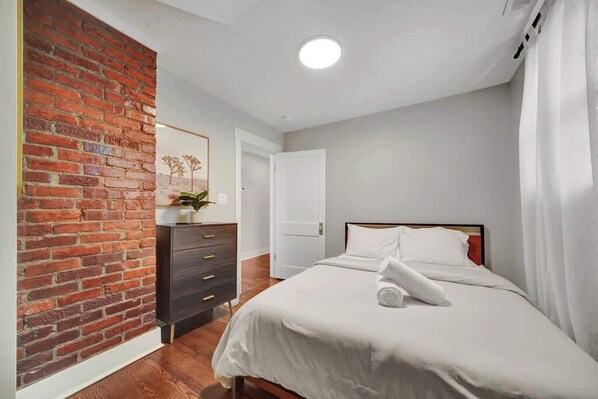 A bedroom with a brick wall, a bed with white bedding, a dresser, and a door.