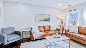 A living room with a brown leather sofa, a grey armchair, a white coffee table, and a painting on the wall. - Spacious unit near Downtown and Bourbon Trail! (Louisville)