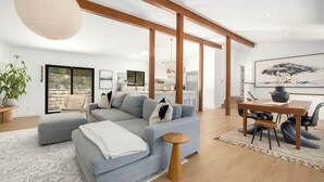 Un salon moderne avec un canapé d’angle, une petite table d’appoint ronde en bois et un coin repas comprenant une table et des chaises en bois.