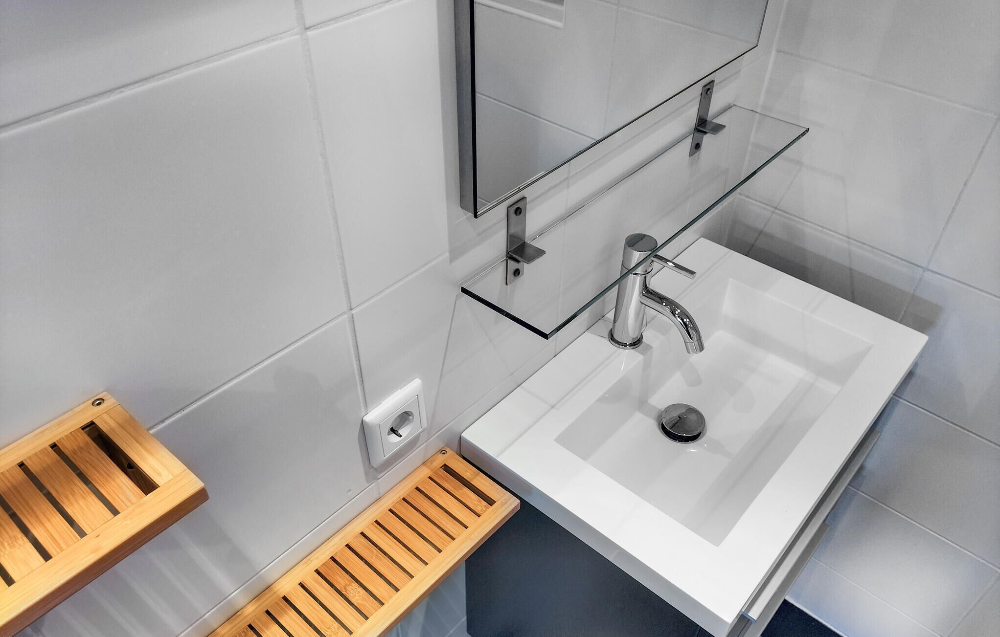 A modern bathroom with a white sink, chrome faucet, and a glass shelf above it. There is a wooden tray under the sink and a mirror on the wall.