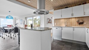 A modern kitchen with white cabinetry, a granite countertop, and a built-in dishwasher. There is a dining area with a table and chairs, and a large window offering a view of the outdoors. - Lovely home in Fur with sauna (Fur)