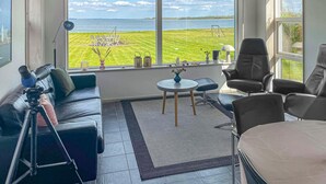 A modern living room with a large window overlooking a grassy field and a body of water. There is a sofa, a chair, a small table with a vase, and a tripod with a camera. - Lovely home in Fur with sauna (Fur)