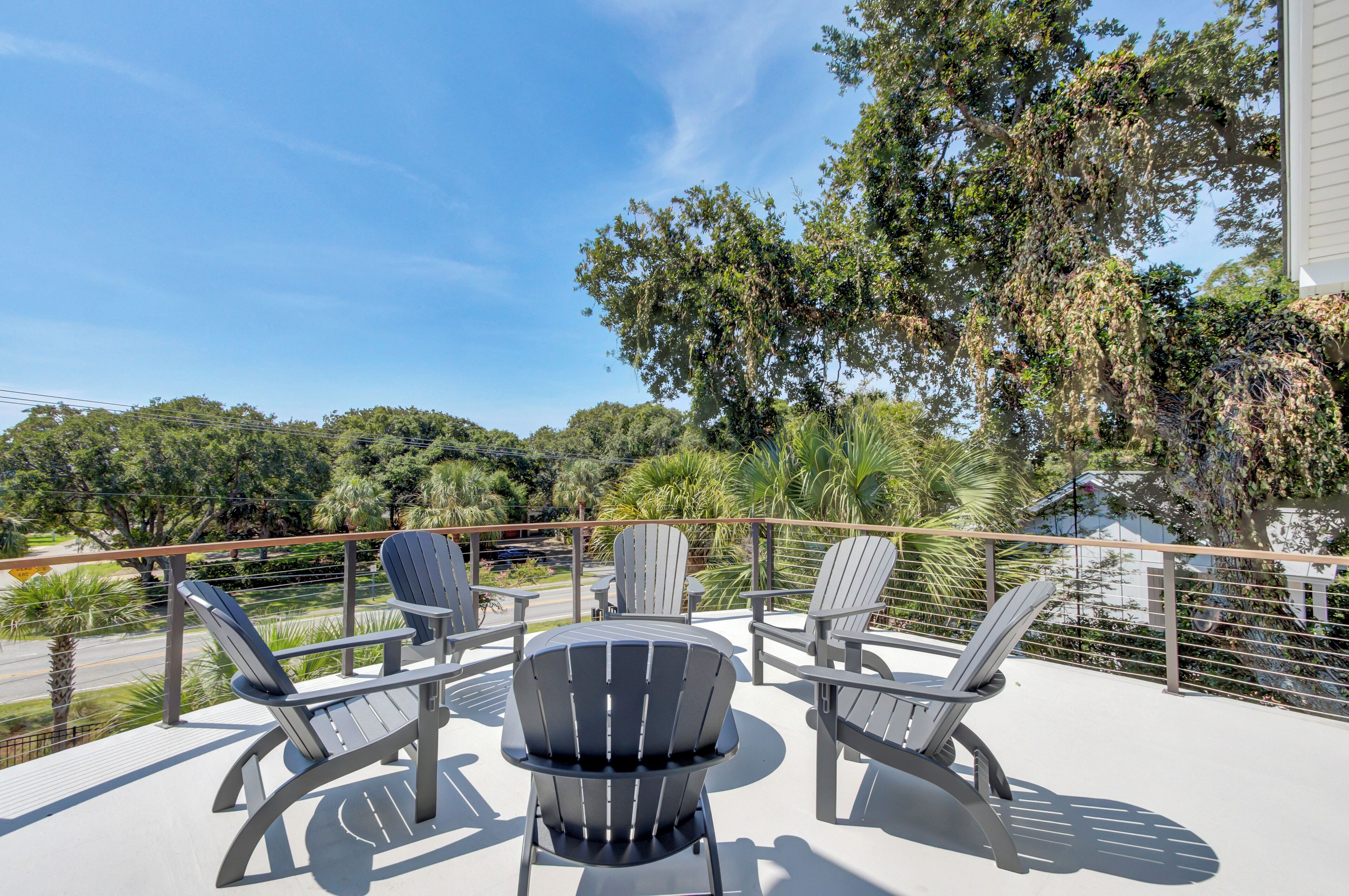 A balcony with Adirondack chairs and a round table, overlooking a lush green landscape and a road.