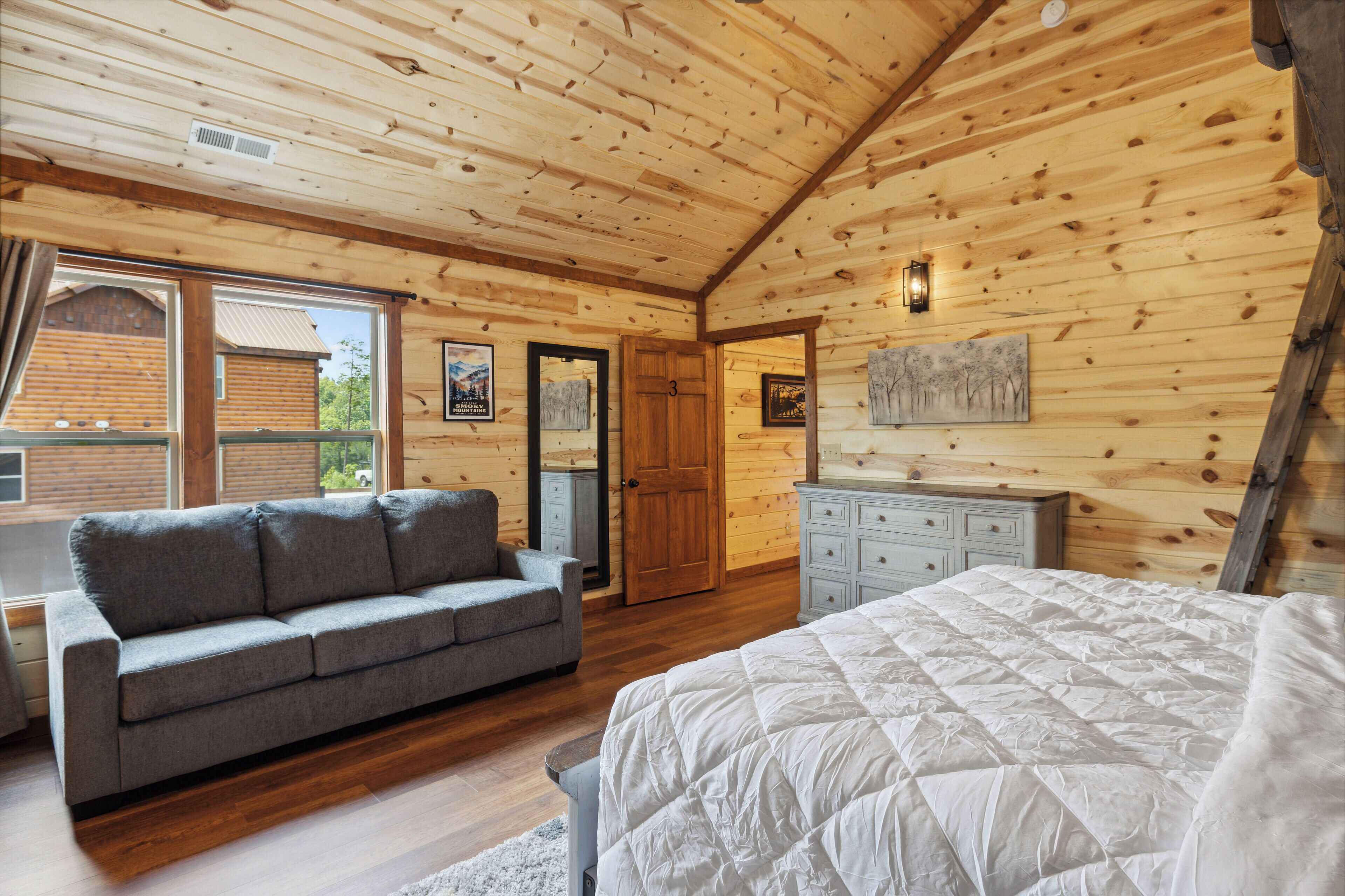 A log cabin bedroom with a gray sofa, a white bed, wooden walls, a dresser, and a door marked with the number 3.