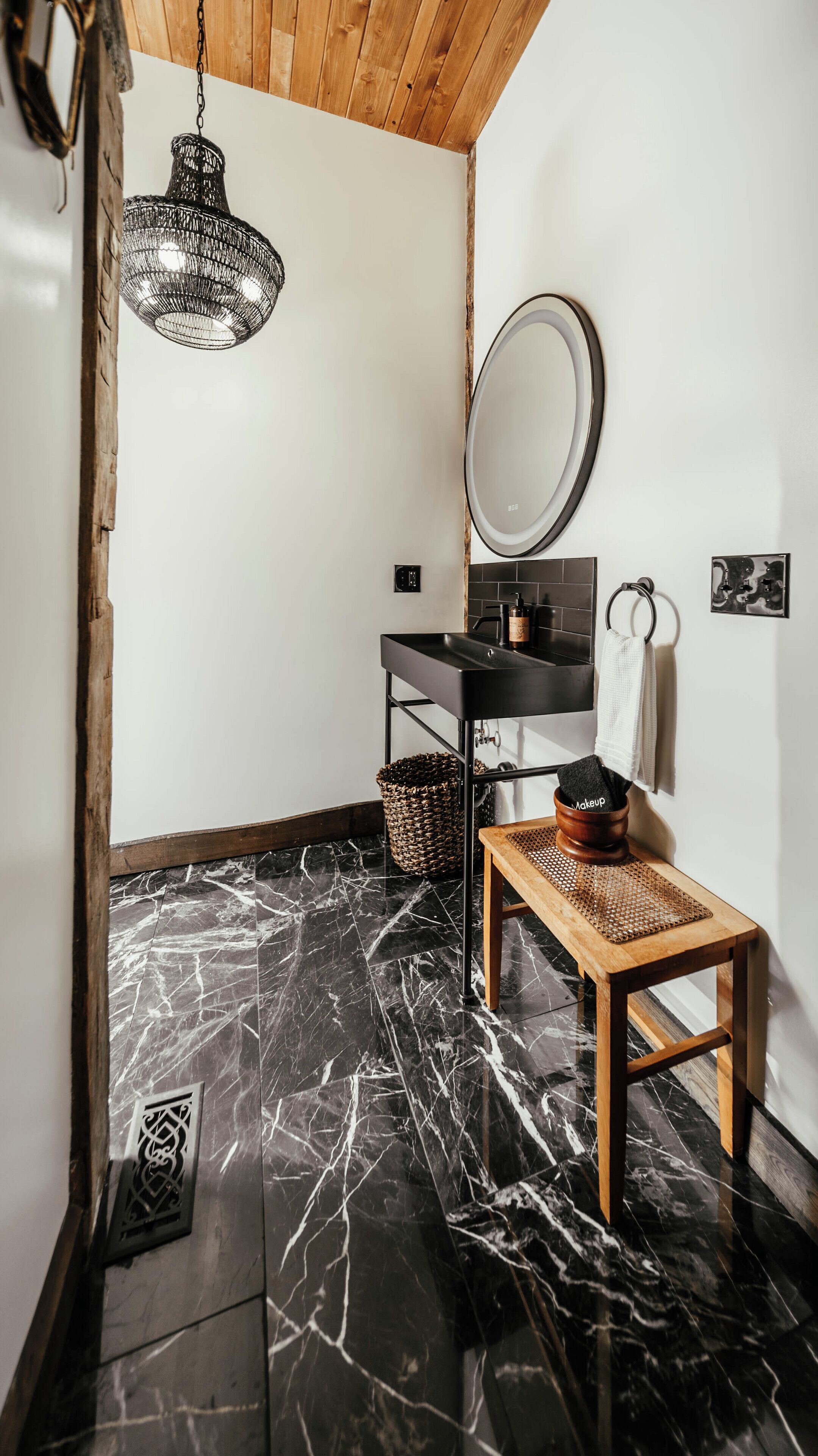A bathroom with a wooden bench, a black marble floor, a hanging light fixture, a mirror, and a sink with a black countertop.