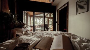 Ein Schlafzimmer mit einem Bett, einem offenen Buch und Blick auf einen Wald durch große Fenster.