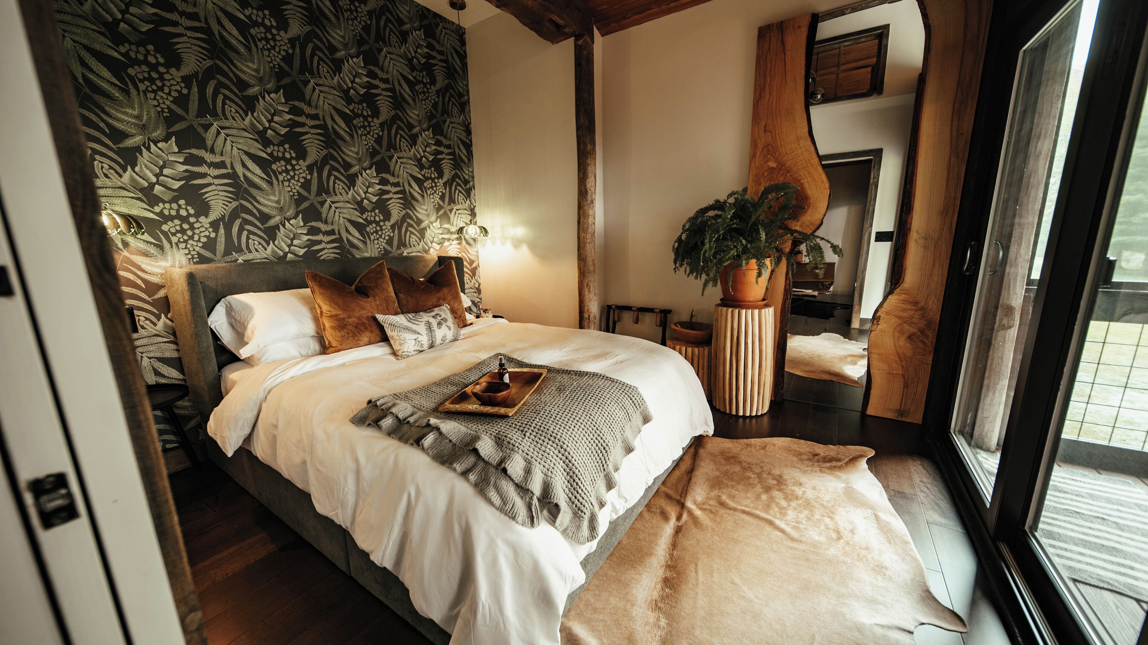 Ein Schlafzimmer mit einem großen Bett, einem Holzkopfende, einer Topfpflanze und Blick ins Freie.