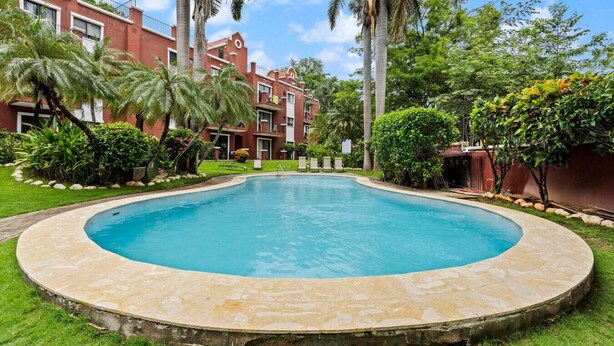 A swimming pool surrounded by lush greenery and a red building with balconies. - Classy Condo in the heart of town (Tamarindo)