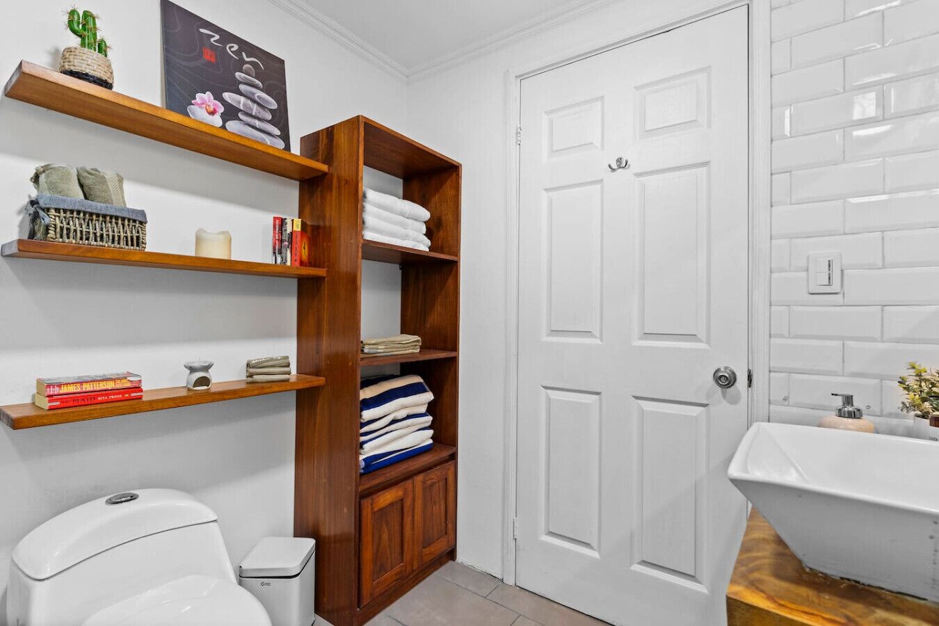 A bathroom with a white toilet, a wooden shelf unit with towels and books, a white door, and a white sink with a mirror.