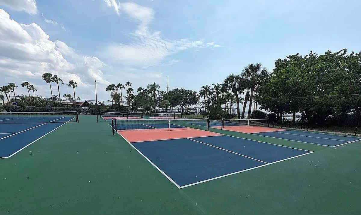 Tenniskenttä, jossa on useita kenttiä, ja sen ympärillä on palmuja ja kirkas taivas.