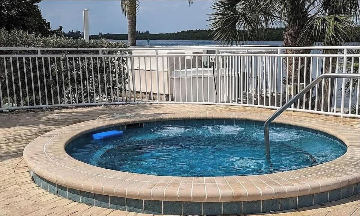 A hot tub with a tiled edge, surrounded by a white metal fence, and a view of a body of water in the background.