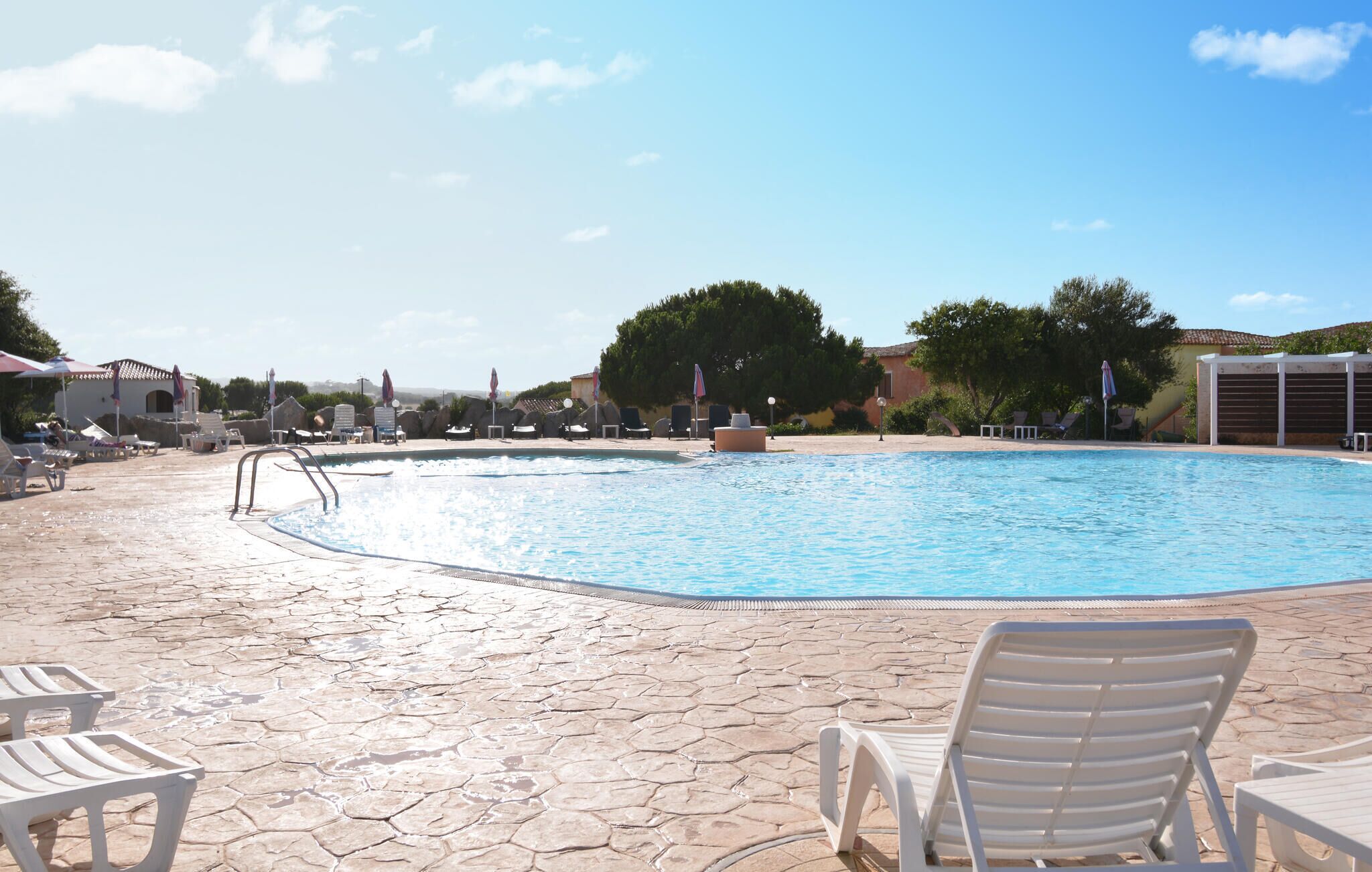 A swimming pool with lounge chairs and umbrellas, surrounded by trees and buildings.