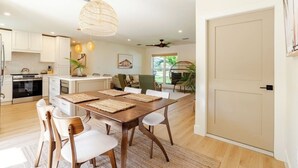 Une cuisine moderne avec des meubles blancs, un coin repas doté d’une table et de chaises en bois, et une grande fenêtre qui laisse entrer la lumière naturelle.