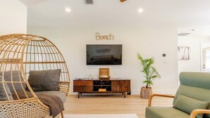 Un salon avec un fauteuil en osier, un canapé vert, un meuble de rangement en bois et un téléviseur fixé au mur.