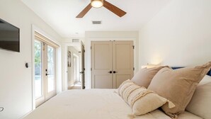 Une chambre à coucher comprenant un lit, un ventilateur de plafond, un téléviseur fixé au mur et une porte donnant sur une autre pièce.