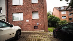 A red brick building with multiple windows, a paved driveway, and two parked cars.