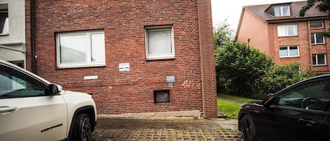 A red brick building with multiple windows, a paved driveway, and two parked cars.