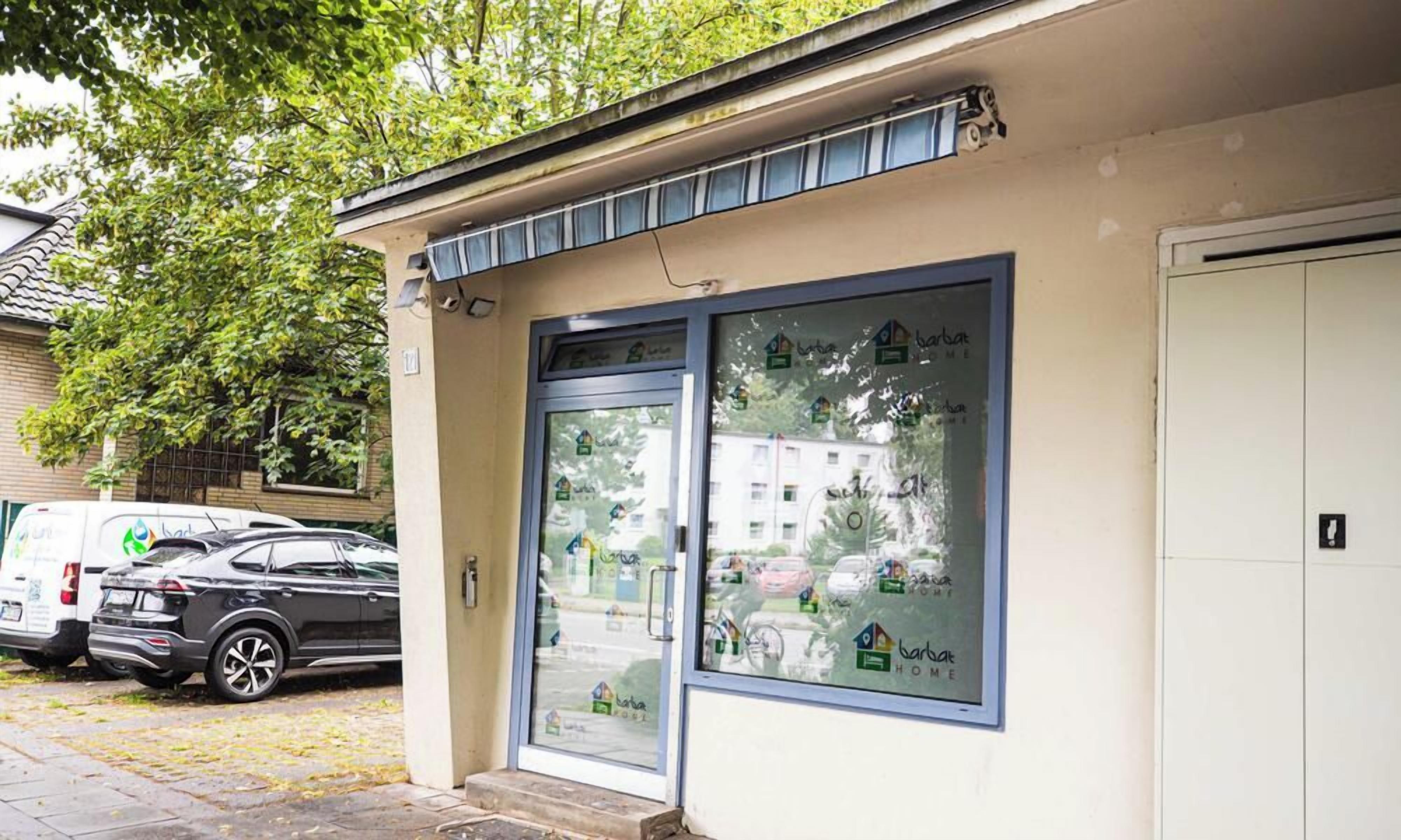 Ein Gebäude mit Glastür, ein Parkplatz mit Autos und ein Baum im Hintergrund.