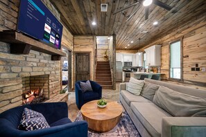 Un salon chaleureux avec une cheminée en pierre, un téléviseur à écran plat fixé au mur, un plafond en bois et un escalier menant à un espace cuisine.