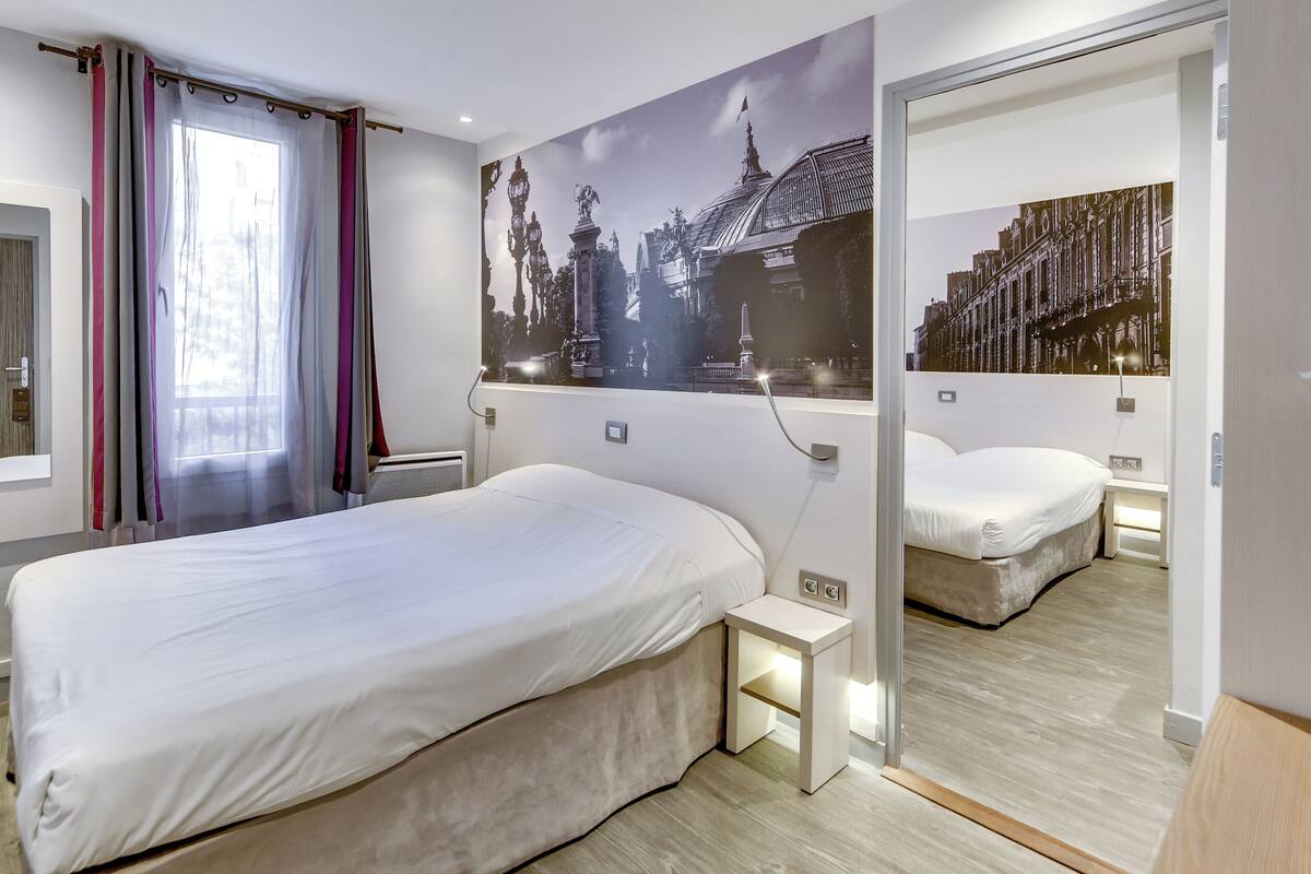 Camera d'albergo con un letto grande, un comodino, uno specchio e un'opera d'arte appesa al muro raffigurante un paesaggio urbano.