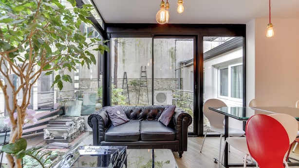 Un salon moderne avec un canapé en cuir noir, une table basse en verre et un fauteuil rouge. Une grande fenêtre offre une vue sur l’extérieur, et une plante est visible au premier plan.