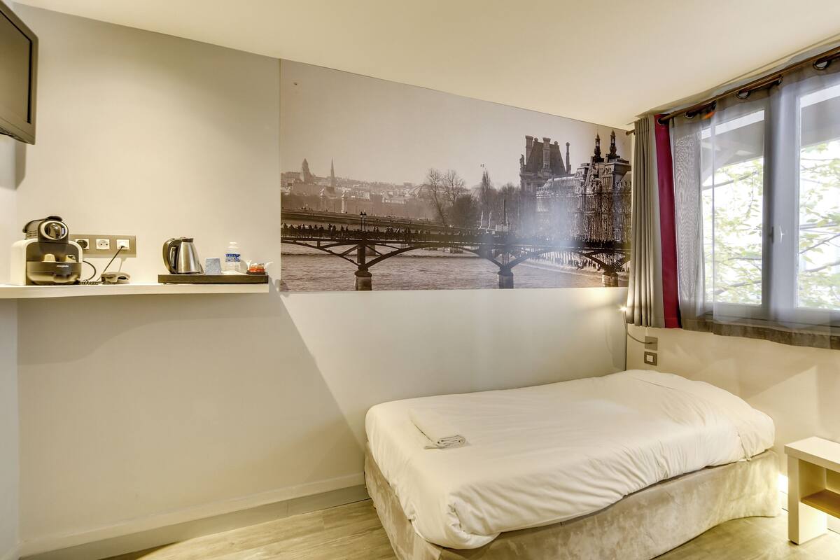 Una camera da letto con un letto, uno scaffale a parete con articoli da toilette, una finestra con tende e un quadro che raffigura una veduta cittadina.