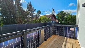 Een houten terras met zonnepanelen, een metalen hek en een uitzicht op een gebouw met rode daken en een klokkentoren in de verte.
