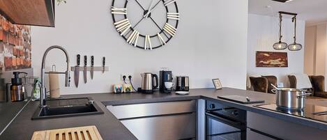 A modern kitchen with a large wall clock, stainless steel appliances, and a wooden cutting board.