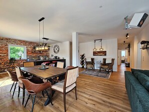Ein geräumiger Wohnbereich mit einem Holzesstisch, Ledersesseln und einem grünen Sofa. Im Hintergrund ist eine Küche mit Backsteinwand und modernen Geräten zu sehen.