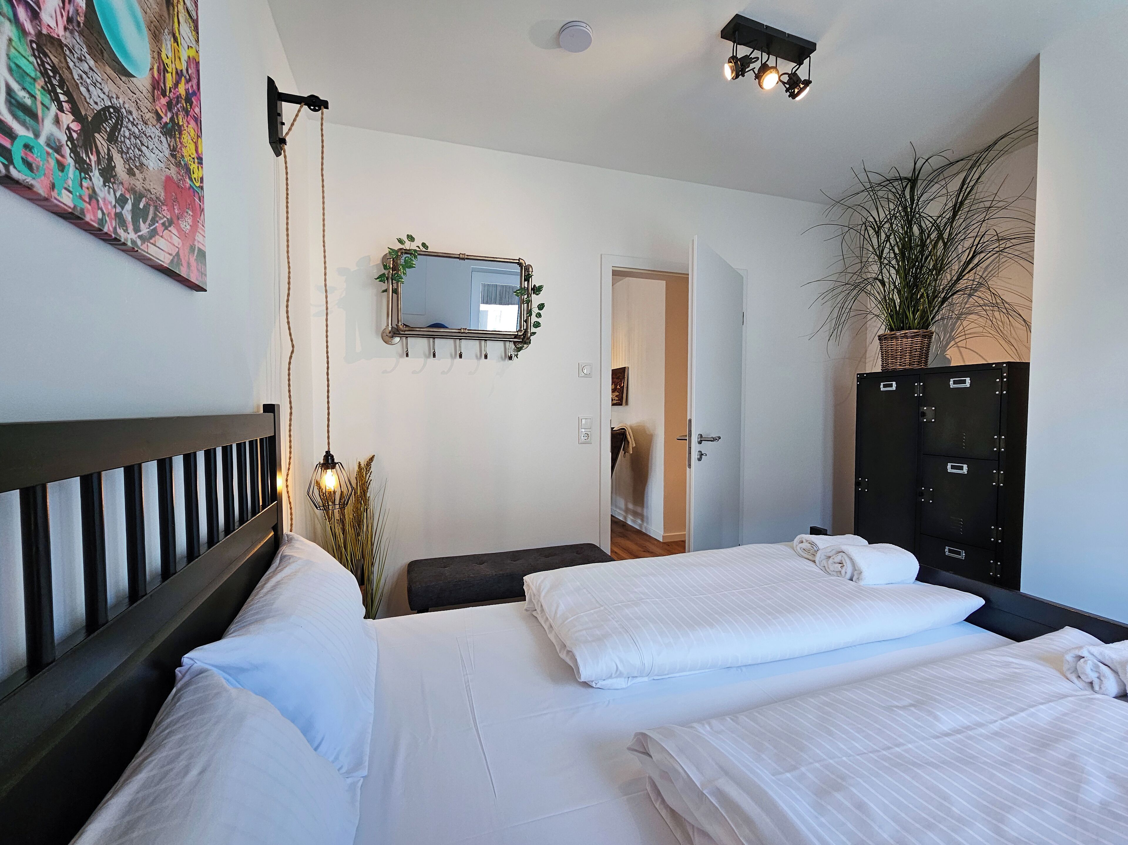 Una habitación de hotel con cama, mesitas de noche, un espejo, una planta en maceta y un armario.