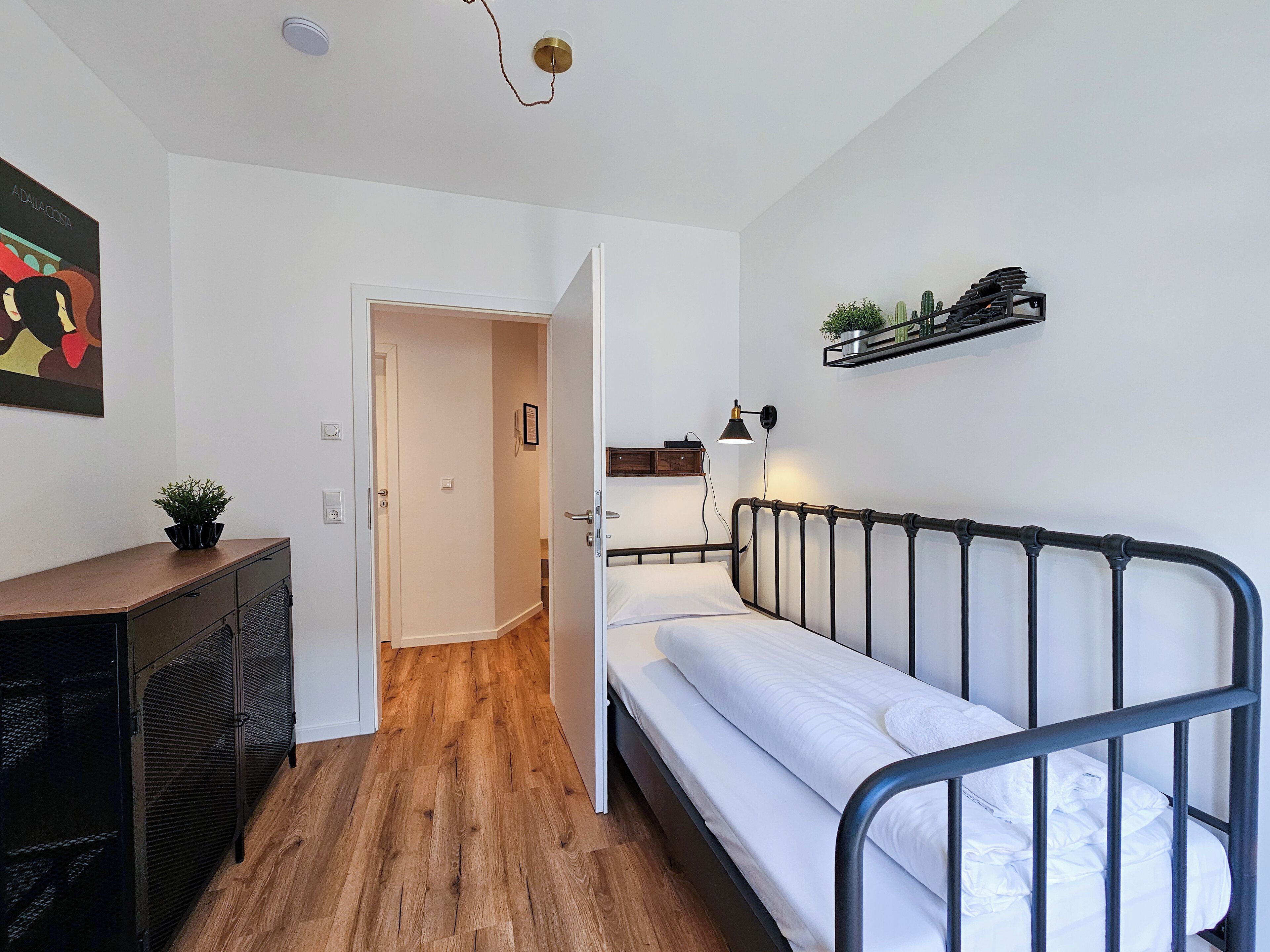 Une chambre à coucher comprenant un lit, une commode, une porte donnant sur une autre pièce et une étagère murale ornée de plantes.