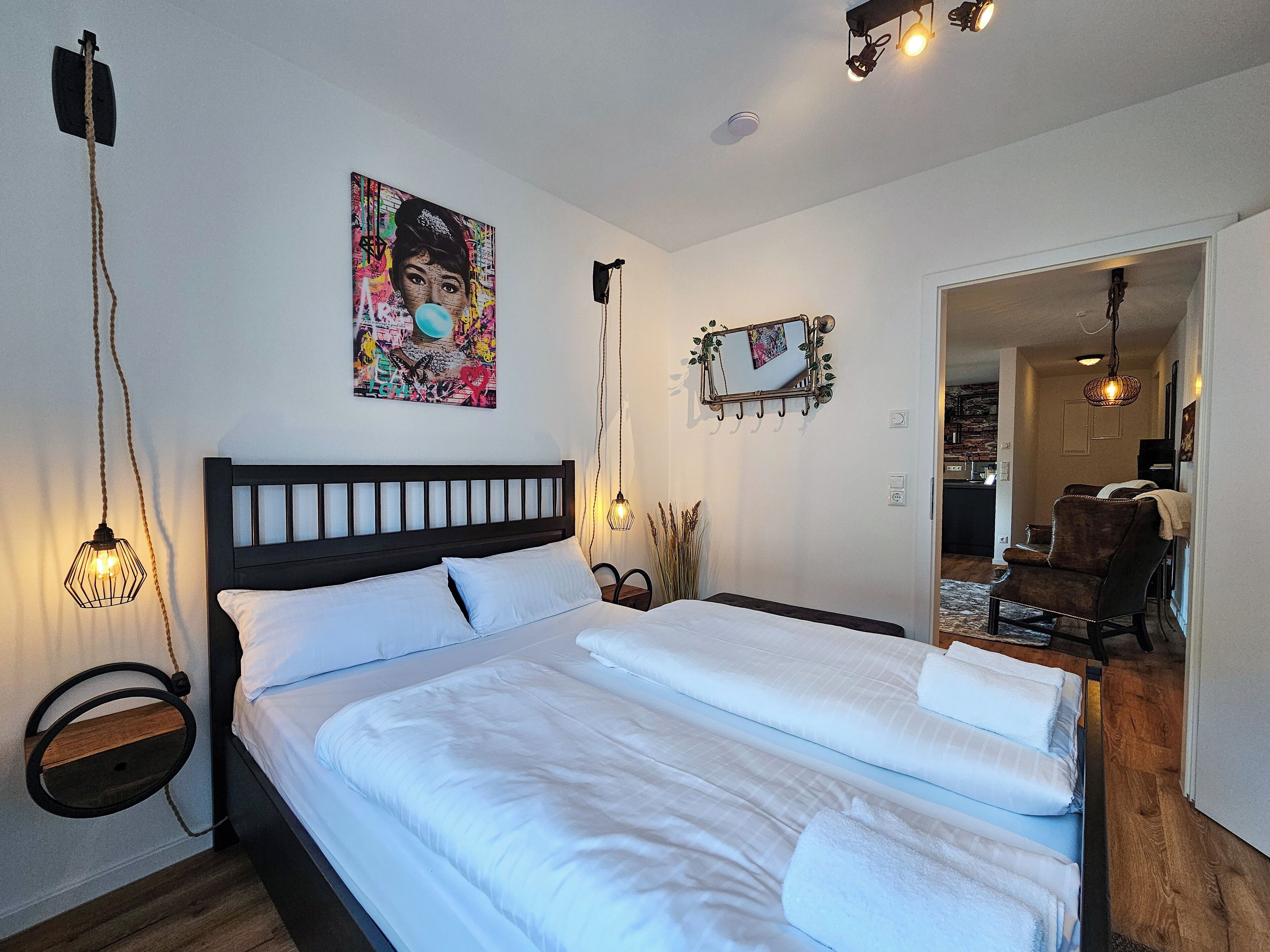 A neatly arranged bedroom with a large bed, a bedside table, a hanging light fixture, and a wall-mounted mirror.