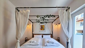 Een hemelbed met witte lakens en een houten frame, twee bedlampen en uitzicht buiten door een groot raam.